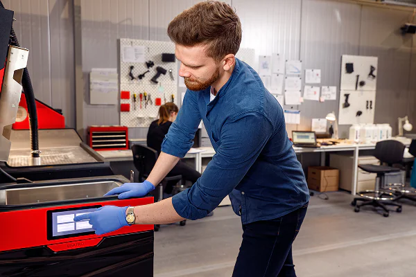 Close-up of hands in blue gloves cleaning a 3D printed part using a wire brush inside a powder handling station.