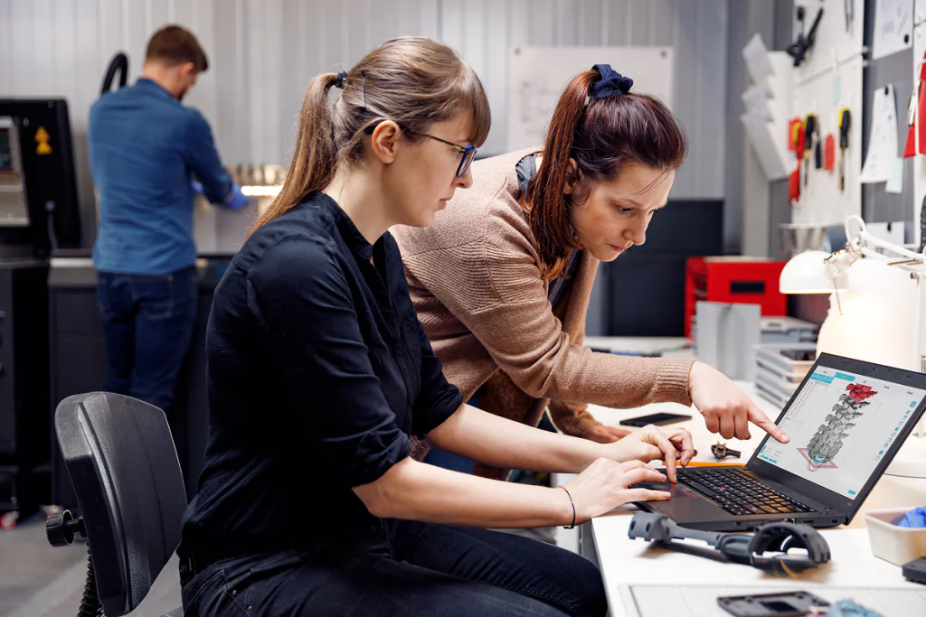 Two women working together on a 3D model displayed on a laptop in a modern workspace, with a man operating 3D printing equipment in the background.